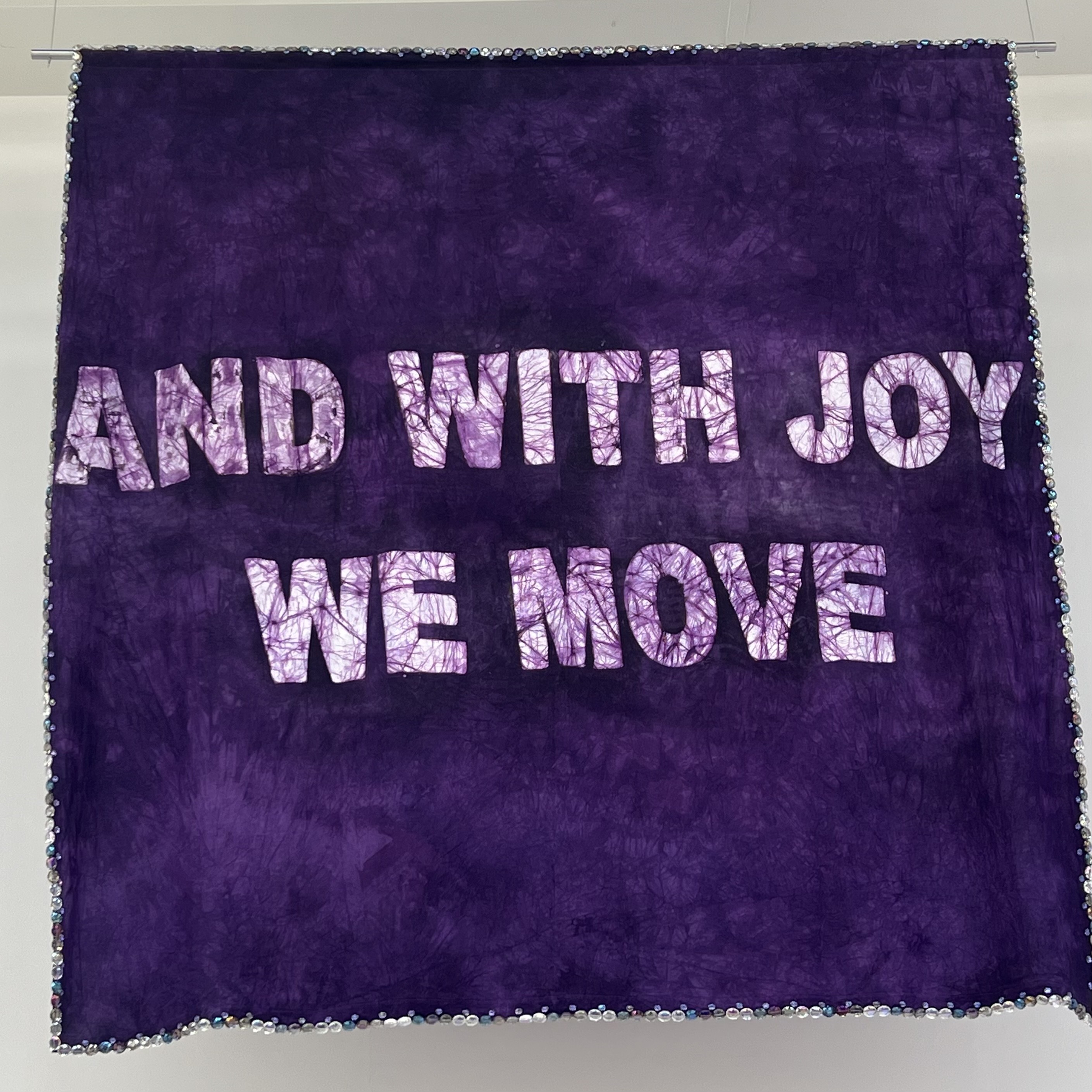 Photo d'une œuvre suspendue : un grand carré violet bordé de perles, avec écrit au milieu 'and with joy we move'